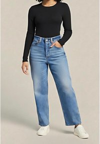 Personne portant un haut noir à manches longues, un jean bleu taille haute avec une main dans la poche et des baskets blanches, debout sur un fond neutre.