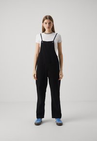 Sorte bomullsloverall med to frontlommer, justerbare stropper, brukt over en hvit T-skjorte, kombinert med blå sneakers mot en enkel bakgrunn.