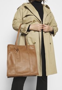 Sac fourre-tout en cuir marron de forme rectangulaire plate, avec deux longues anses et une petite étiquette. Le modèle porte un trench beige et un haut noir.