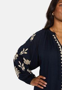 Marineblauwe blouse met lange, volumineuze mouwen, voorzien van gouden bloemenborduursel en een keyhole-halslijn. Zachte stof met elastische boorden en decoratieve afwerking.