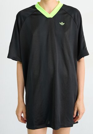 Osoba nosząca czarną sportową koszulkę oversize z neonowo zielonym kołnierzem w kształcie litery V i małym logo na lewym piersi.