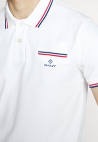 Biała koszulka polo wykonana z teksturowanej tkaniny, z dwoma guzikami i kieszenią na piersi z czerwono-niebieskimi akcentami. Wyświetlone logo GANT.