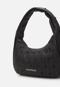 Sac à main Valentino noir avec un motif en losanges rembourré, des clous pyramides noirs, une anse rembourrée et un logo argenté sur le devant, le tout sur fond blanc.
