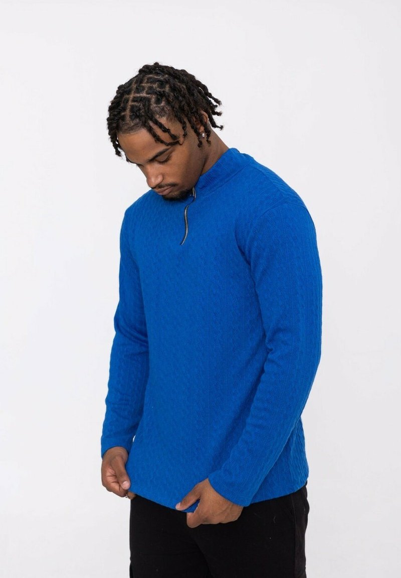 Pull-over bleu texturé à manches longues avec une demi-fermeture éclair, présentant un motif en losanges et des poignets ajustés, porté avec un pantalon noir.