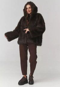 Brązowa kurtka z sztucznego futra z kapturem i szerokimi rękawami, z zamkiem błyskawicznym z przodu. W połączeniu z ciemnobrązowymi spodniami i masywnymi czarnymi butami na platformie.