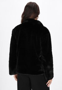 Donna con lunghi capelli scuri che indossa una giacca in pelliccia sintetica nera, pantaloni neri e orecchini a cerchio dorati, vista di spalle.