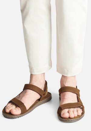 Piedi con sandali marroni aperti con cinturini in Velcro, abbinati a pantaloni corti color crema su uno sfondo chiaro e uniforme.