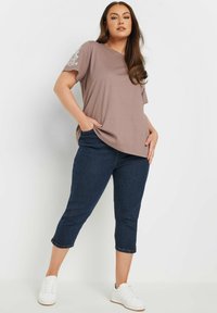 Ljus taupe kortärmad topp med blommig brodyr på vänster axel, kombinerad med mörka denim capribyxor och vita sneakers.