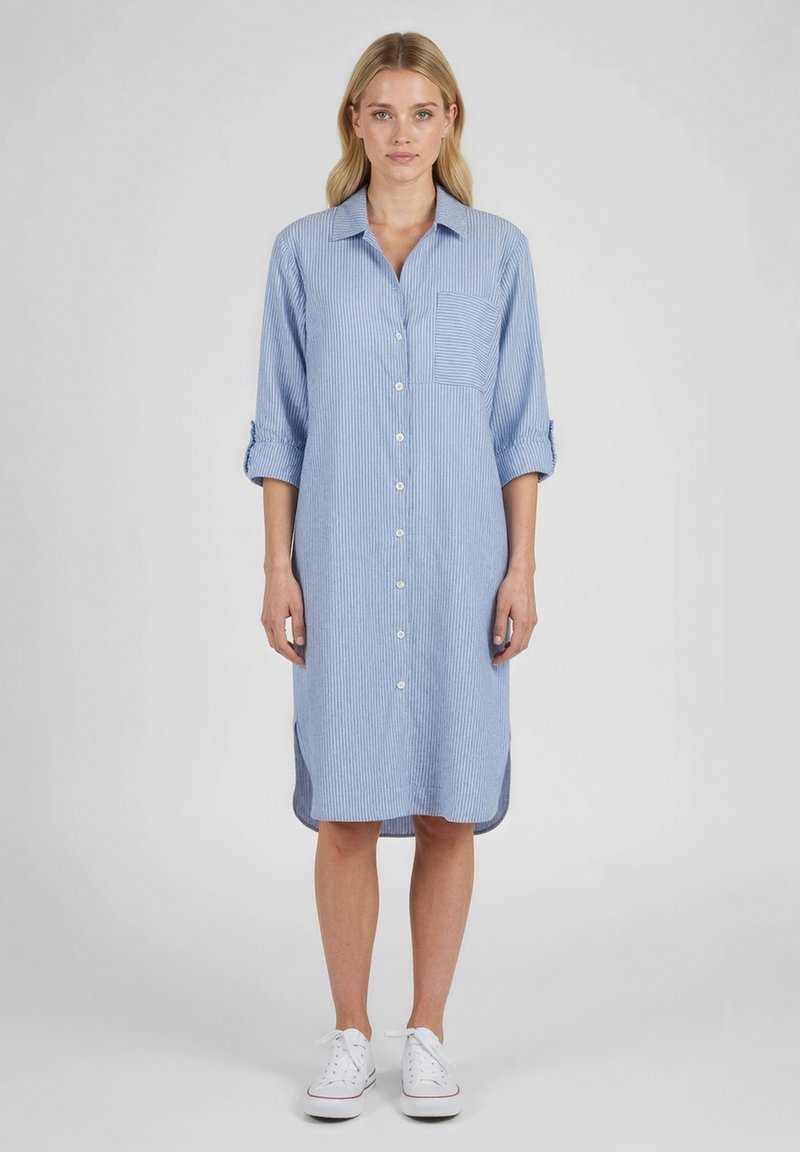 Femme debout portant une robe chemise bleu clair, boutonnée, longueur genou, manches retroussées et baskets blanches, sur un fond gris.