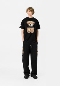 Camiseta de algodón negra con un gráfico de oso de peluche, combinada con pantalones de chándal negros con acentos de oso de peluche. Diseño casual y de corte relajado.