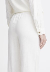Weiße Leinenhose mit elastischem Bund und Seitentaschen. Leicht strukturierten Stoff, mit einem detailreichen Bundabschluss und einem Knopfakzent.