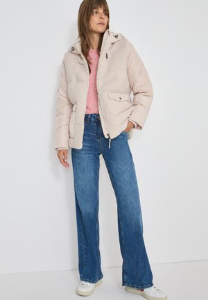 Giacca a vento beige con parte anteriore testurizzata, chiusura con zip e cappuccio, abbinata a jeans svasati azzurri e sneakers bianche, su uno sfondo neutro.