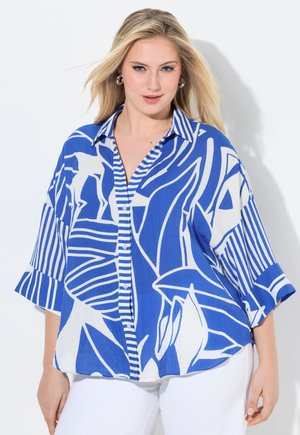 Lyshåret kvinde iført en løs blå og hvid mønstret skjorte med knapper og brede ærmer, kombineret med hvide bukser, mod en hvid baggrund.