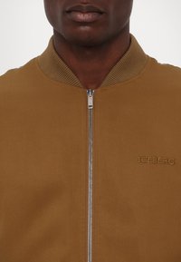 Barna bombázódzseki bordás gallérral, elülső cipzárral és hímzett "ICEBERG" logóval. Sima szövettextúra, testhezálló fazon.