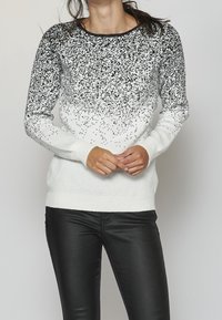 Pull en tricot avec un motif dégradé, comportant des taches noires sur une base blanche. Col rond, manches longues, et poignets et ourlet côtelés.