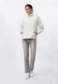 Witte fleecetruiker met een hoge kraag, rits en twee knopen bij de hals. Gekleed met lichtgrijze jeans en witte sneakers.