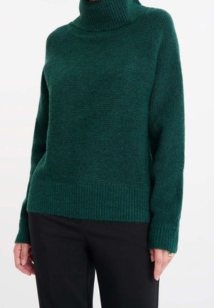 Pull à col roulé côtelé vert foncé porté avec un pantalon noir, montrant des manches longues et une coupe décontractée sur une personne.