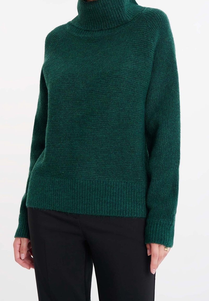 Pull à col roulé côtelé vert foncé porté avec un pantalon noir, montrant des manches longues et une coupe décontractée sur une personne.