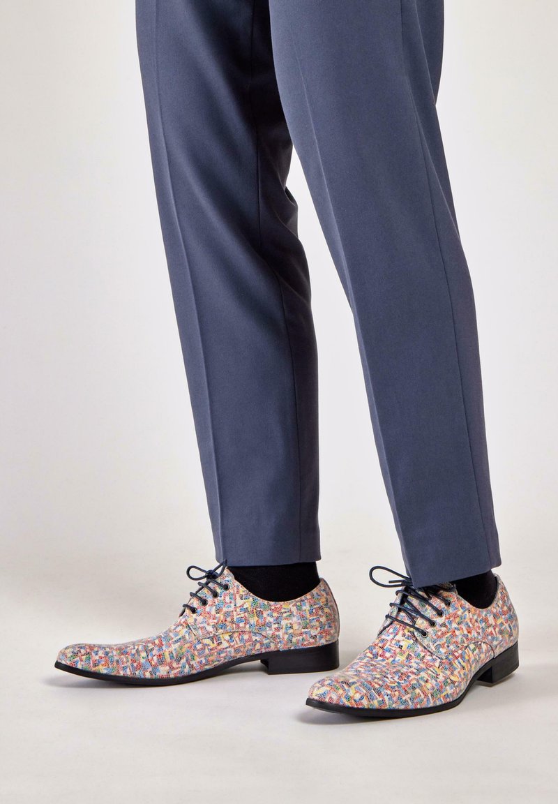 Multicolored patterned dress shoes made of smooth leather, featuring black laces and a low square heel, paired with tailored navy trousers.