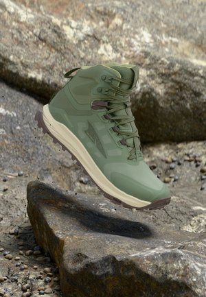 Botte de randonnée imperméable verte avec semelle robuste suspendue au-dessus d'un terrain rocheux, mettant en avant la durabilité en plein air et l'adhérence.