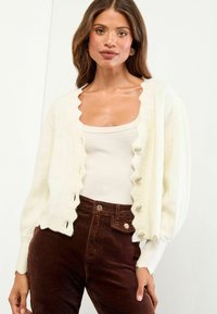 Egy nő krémszínű, fodros szélű kardigánt visel egy bézs ujjatlan felső felett, sötétbarna kordbársony nadrággal, egy sima fehér háttér előtt állva.