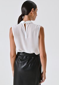Vrouw met donker haar in een lage paardenstaart, gekleed in een mouwloze witte blouse met een knoopsluiting op de rug en een zwarte leren rok, met haar gezicht naar de achterkant gericht.
