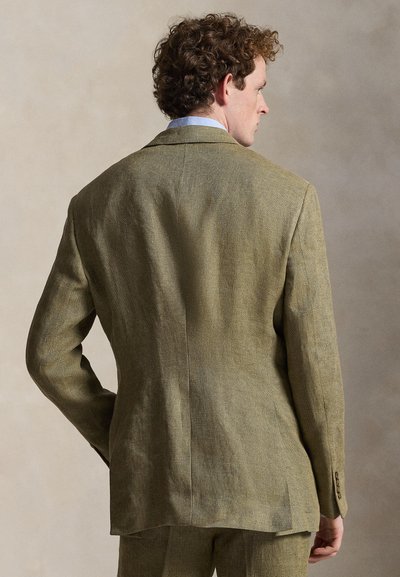 Mann mit lockigem Haar, der einen strukturierten olivgrünen Anzug und ein hellblaues Hemd trägt, steht mit dem Rücken zu einer neutralen Hintergrund.