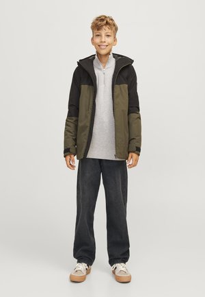 Veste verte et noire avec une capuche, chemise zippée grise en dessous, associée à un jean sombre ample et des baskets beiges. Texture douce, design décontracté.