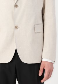 Blazer in lino beige con due bottoni, rever a V, tasca sul petto singola, tasca laterale piccola e un taglio pulito all'orlo. Pantaloni neri visibili.