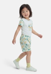 Μικρό παιδί που περπατά, φοράει λευκό πουκάμισο με το κείμενο "ΝΑ ΕΙΣΑΙ ΑΓΡΙΟΣ" και σορτς με σχέδια ζώων και φυτών, λευκά αθλητικά παπούτσια και κίτρινες κάλτσες.