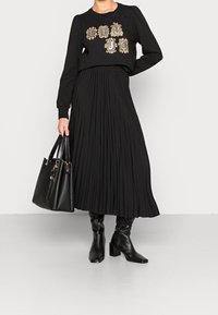 Sudadera negra de manga larga con bordado dorado, falda midi plisada negra y botas negras de tacón a la altura de la rodilla. Bolso negro.