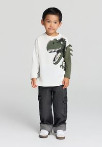 Biała koszula z długim rękawem z grafiką dinozaura z przodu i oliwkowym rękawem. Zestaw z ciemnymi spodniami cargo i białymi trampkami.