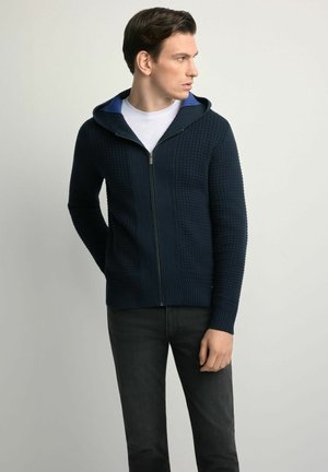 Granatowa dzianinowa bluza z kapturem o strukturalnym wzorze i kontrastującym, niebieskim wnętrzu. Posiada zamek błyskawiczny oraz ściągacze na rękawach i u dołu.
