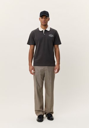 Uomo che indossa un berretto nero, una polo scura con colletto chiaro, pantaloni beige con motivo e scarpe nere, in piedi davanti a uno sfondo chiaro e uniforme.