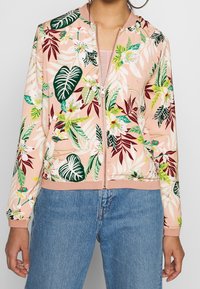Chaqueta bomber floral con base de color rosa claro, que presenta hojas y flores en tonos verdes y burdeos, cierre con cremallera y puños y dobladillo acanalados.
