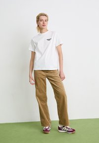 Person steht auf grünem Boden und trägt ein weißes T-Shirt mit Druck, braune Cordhosen und burgunderfarbene und weiße Sneaker vor einer weißen Wand.