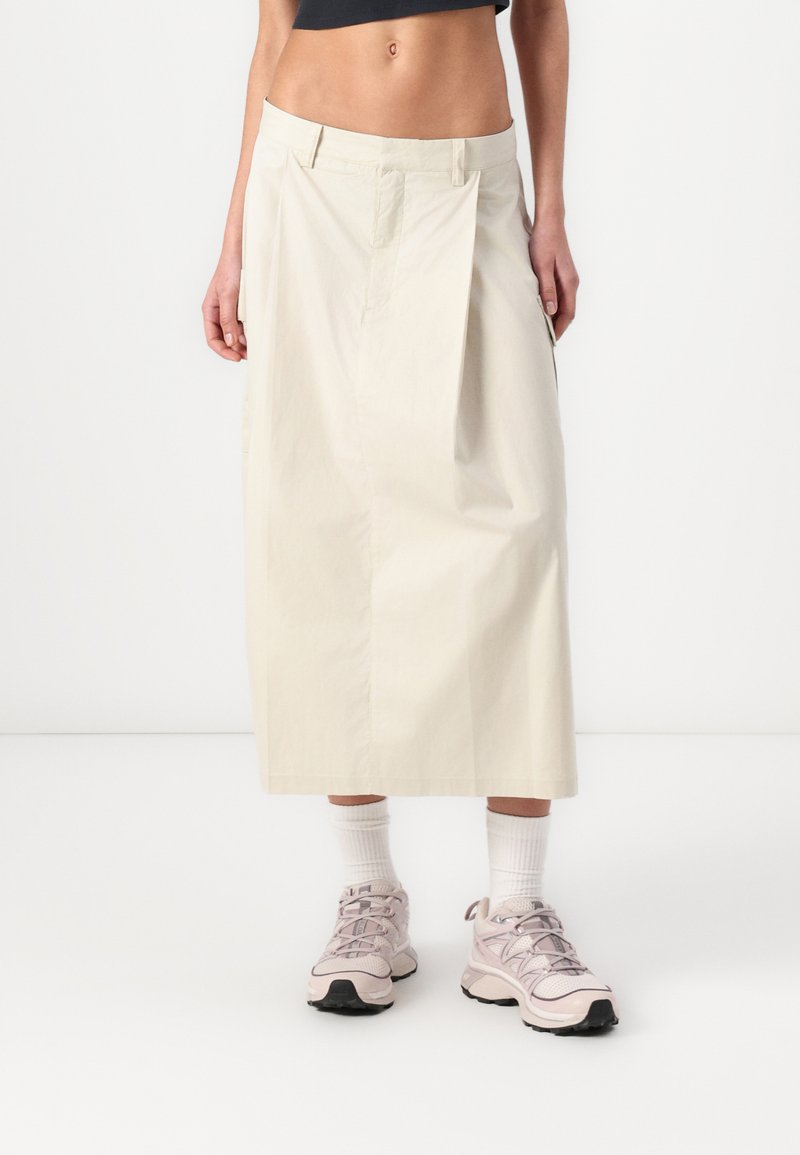 Light beige ankle-length skirt made of smooth fabric, featuring a pleated design and side pockets, paired with pale pink athletic shoes.