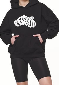 Felpa nera con cappuccio e logo astratto bianco, dotata di tasca a canguro e polsini a coste. Abbinata a pantaloni ciclistici neri.