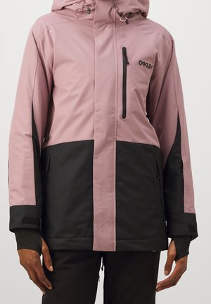 Frau trägt eine zweifarbige rosa-schwarze Kapuzenjacke mit Reißverschlüssen an Brust- und Seitentaschen sowie Armbändern an den Ärmeln.