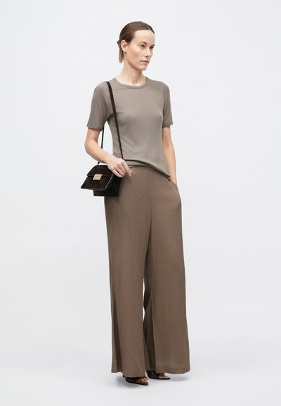 Sieviete ar gaiši brūnu īsroku topu un platajiem brūnajiem biksēm, turot mazu melnu somiņu ar zelta aizdari uz vienmērīga fona.