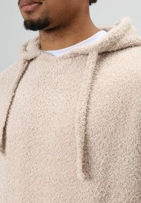 Beige fleece hoodie met een gestructureerde, zachte stof. Voorzien van een trekkoord bij de hals en een eenvoudig ontwerp, met de nadruk op warmte en comfort.