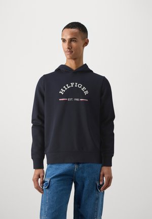 Felpa blu navy con cappuccio e coulisse, dotata di tasca a marsupio frontale. Presenta la scritta "HILFIGER EST. 1985" in bianco con accenti rossi. Tessuto morbido.