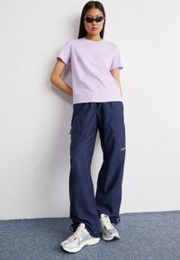 T-shirt a maniche corte di colore viola chiaro con logo, abbinato a pantaloni cargo blu navy con tasche e logo, e sneaker sportive argentate.