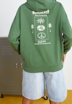 Person trägt grüne Quiksilver Surf Company Kapuzenjacke mit Grafik von Palmen, Globus und Friedenstaube, kombiniert mit hellen Jeansshorts.