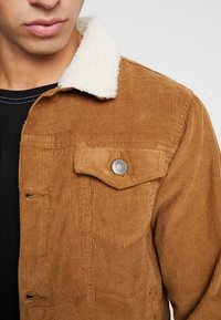 Brązowa kurtka ze sztruksu z białym kołnierzem z merynosów, wyposażona w zapinaną na guziki kieszeń na piersi oraz prążkowaną fakturę. Pod spodem czarna koszulka.