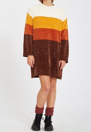 Kvinde iført en lang strikket sweaterkjole med hvide, gule, orange og brune vandrette striber, kombineret med sorte støvler og røde sokker.