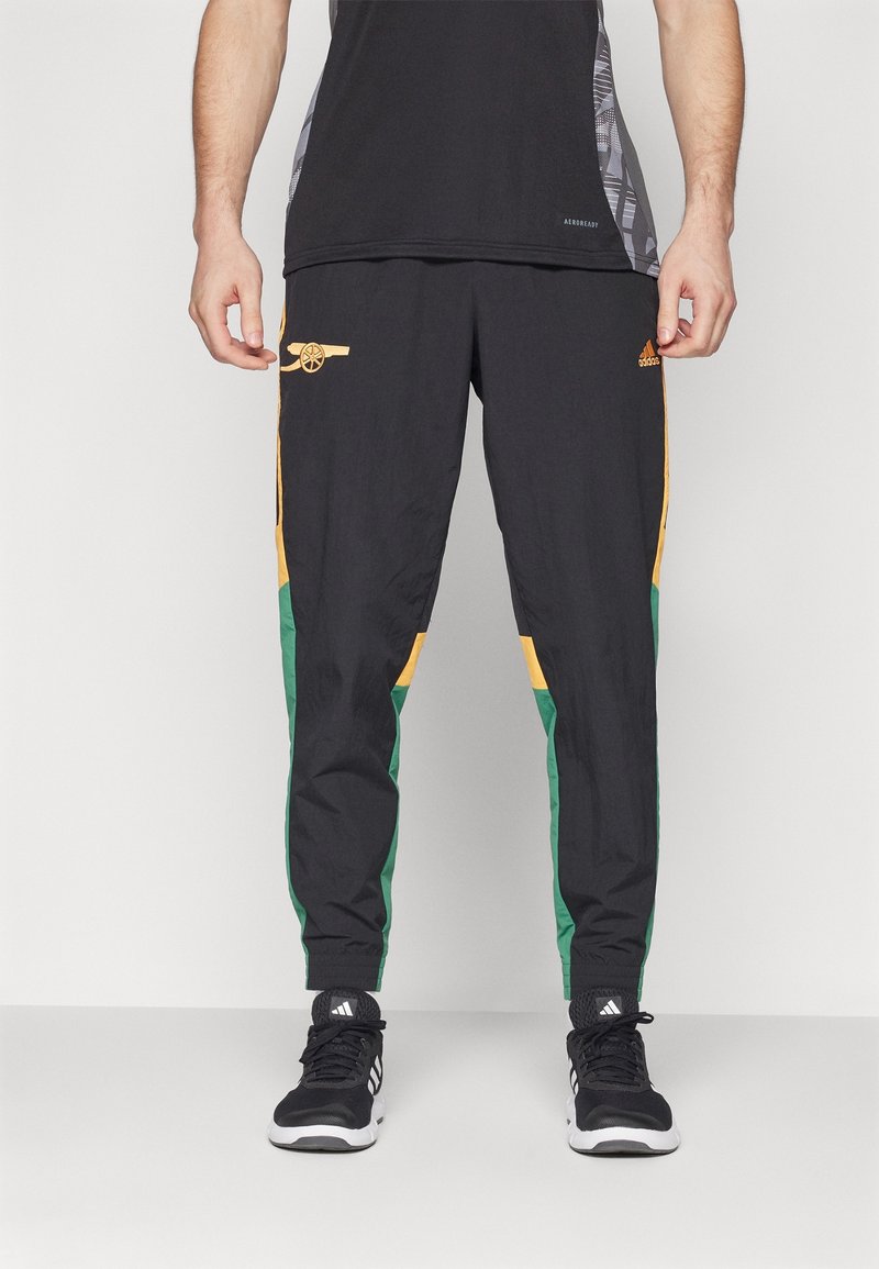 Homme portant un pantalon de sport Adidas noir, vert et jaune avec le logo du club de football Arsenal et des baskets Adidas noires, debout devant un fond uni.