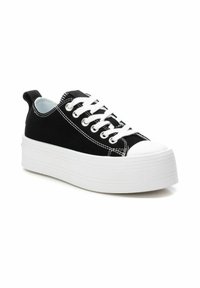 Zapatillas de lona negras con una suela plateada gruesa blanca, cordones blancos y costuras en contraste. Cuenta con una lengüeta en el talón.