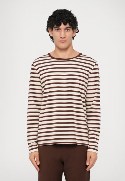 Young man with curly dark hair wearing a long-sleeve white shirt with horizontal brown stripes and solid brown pants, standing against a white background.