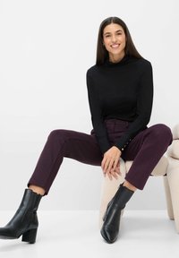 Schwarzer Rollkragenpullover, burgunderfarbene, strukturierte, schmal geschnittene Hose und schwarze Knöchelstiefel mit Reißverschluss. Modell sitzt auf einem hellen Stuhl.